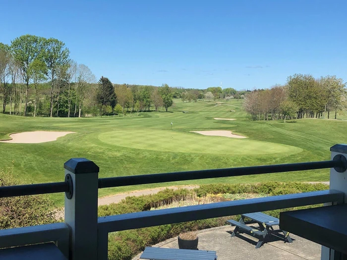 Udsigt fra en balkon over en golfbane med green, sandbunkers, træer og bænke på en solrig dag.