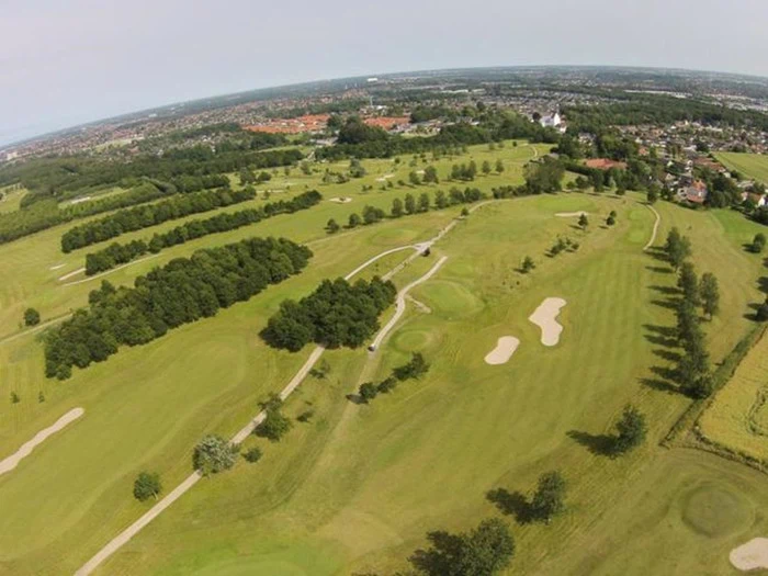 Luftfoto af en stor golfbane med grønne fairways, sandbunkers, træer og omkringliggende bygninger under en klar himmel.