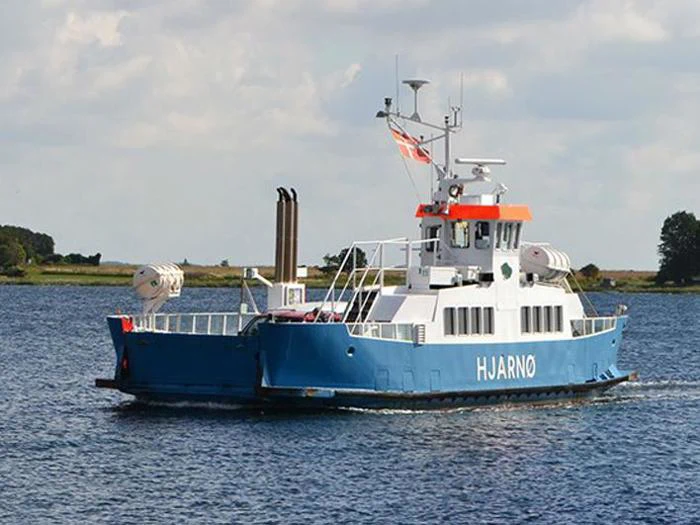 En blå og hvid færge med navnet HJARNØ sejler på roligt vand med træer og land synlige i baggrunden under en delvist overskyet himmel.