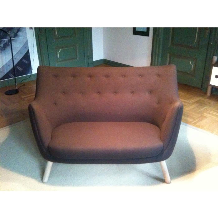 En moderne brun to-personers sofa med knappet ryglæn og lyse træben, placeret på et lyst tæppe i et rum med grønne døre og trægulve.