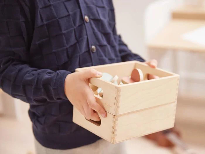 Et barn i en marineblå sweater holder en lys trækasse fyldt med pastelfarvet trælegetøj.