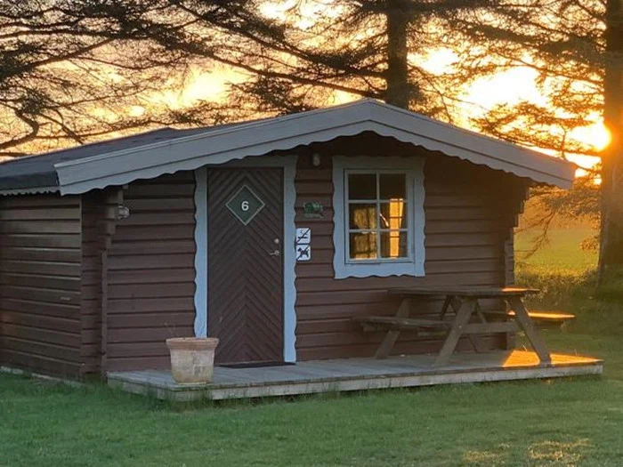 En lille træhytte med veranda og picnicbord ligger mellem træerne i solnedgangen og kaster et varmt, gyldent lys over stedet.