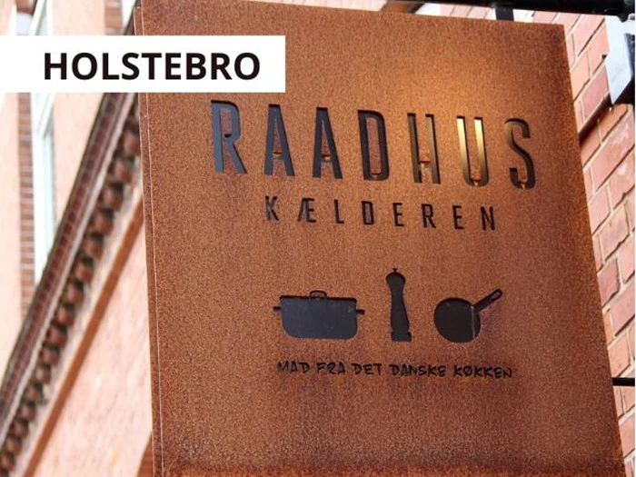 Et rustfarvet metalskilt med teksten RAADHUS KÆLDEREN med køkkenredskabsikoner og dansk tekst nedenunder er monteret på en murstensbygning. HOLSTEBRO står i en hvid boks over skiltet.