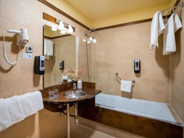 Et rent hotelbadeværelse med spejl, håndvask, hårtørrer, håndklædeholder, badekar, sæbedispensere og foldede hvide håndklæder, der hænger og er udstillet.