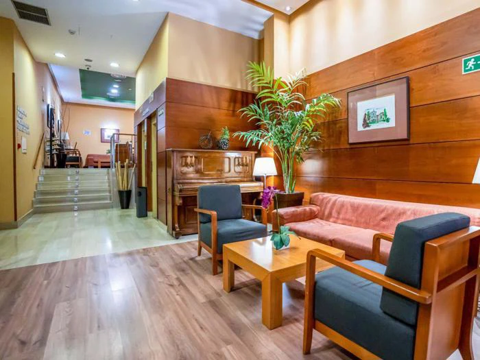 En hyggelig lobby med træmøbler, en sofa, to lænestole, et sofabord, et klaver, potteplanter og en trappe, der fører til et andet område. Varm belysning og vægpaneler af træ.