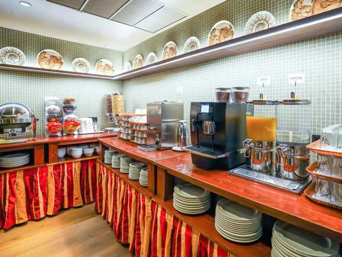 Morgenmadsbuffet med tallerkener, korndispensere, juicemaskine, kaffemaskine, skåle, rødstribede bordplader og dekorative tallerkener på en flisebelagt væg.