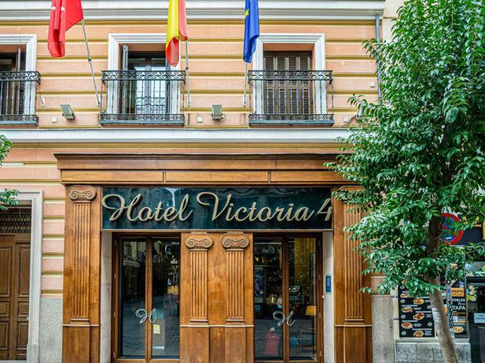 Indgang til Hotel Victoria 4 med store trædøre, søjler og tre balkoner over; flag hængende over indgangen og træer på hver side.