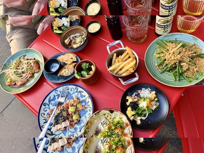 Et rødt bord med forskellige tallerkener med mad, herunder pommes frites, dumplings, stegt tun, grønne bønner, risretter, dypsaucer og drikkevarer, med to personer siddende.
