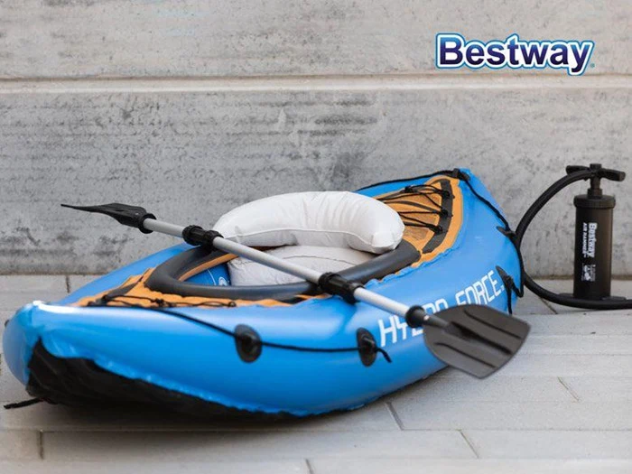 En blå oppustelig kajak med sort pagaj og luftpumpe er placeret på et gråt flisegulv ved siden af en betonvæg. Ordet Bestway står i øverste højre hjørne.