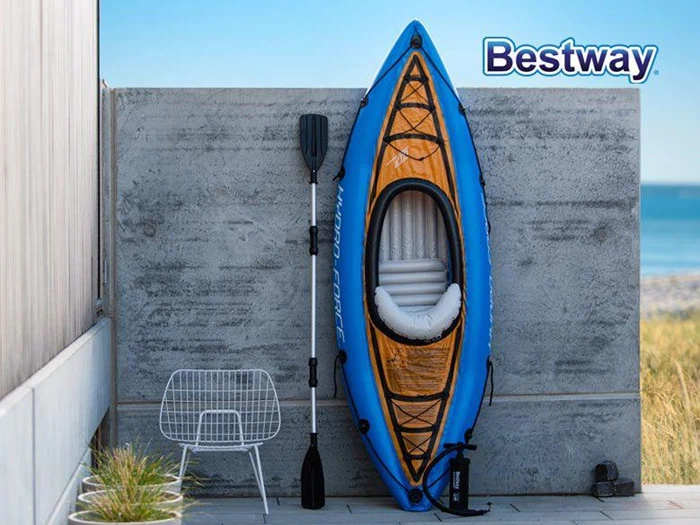 En blå oppustelig kajak med pagaj læner sig op ad en betonvæg i nærheden af en stålstol. Bestway-logoet ses i øverste højre hjørne. Havet og græsset er synligt i baggrunden.