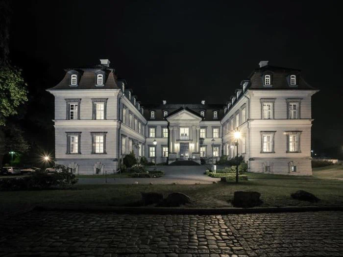 Et stort, elegant palæ med mange oplyste vinduer, set om natten. Bygningen har to fløje, symmetrisk arkitektur og en central indgang med en brostensbelagt sti foran.