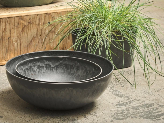 To indlejrede, grå keramikskåle står på en struktureret betonoverflade i nærheden af en potteplante med lange, tynde, grønne blade. En trækasse står i baggrunden.
