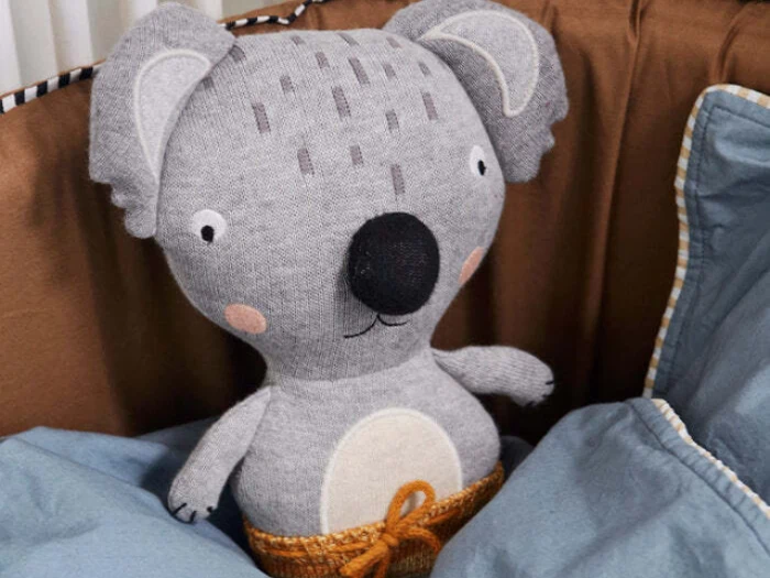 En udstoppet koala med en stor sort næse, rosenrøde kinder og gråmønstret stof sidder blandt blå tæpper og en brun pude.