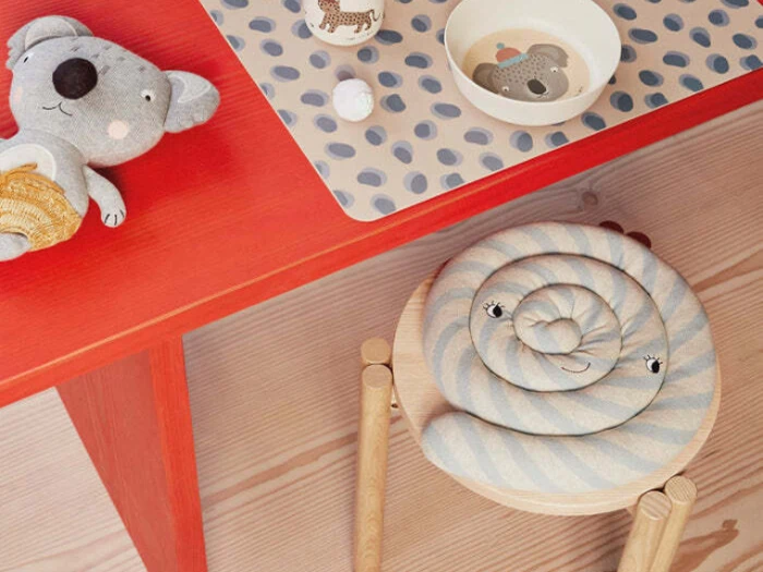 Et børneværelse med et rødt bord, en udstoppet koala, en mønstret måtte, en skål og en træskammel med en blå og hvid spiralformet pude med et smilende ansigt.