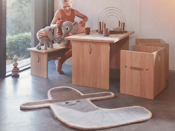 Et barn leger ved et træbord med udstoppede elefanter og legetøj i nærheden af et vindue. Et smilende, abstrakt gråt tæppe ligger på gulvet.