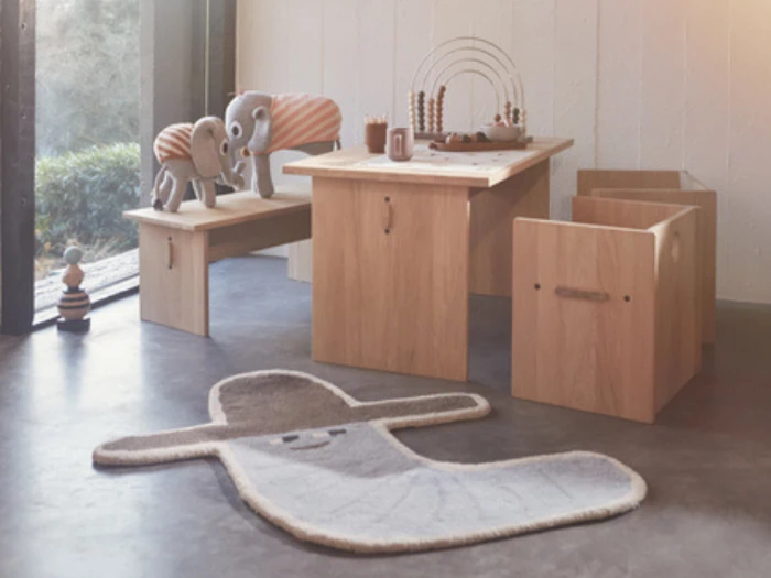 Et hyggeligt legeområde med træbord, stole, en bænk med udstoppede elefanter, perlelegetøj og et tæppe formet som en smilende hval. Sollyset strømmer ind gennem et stort vindue.