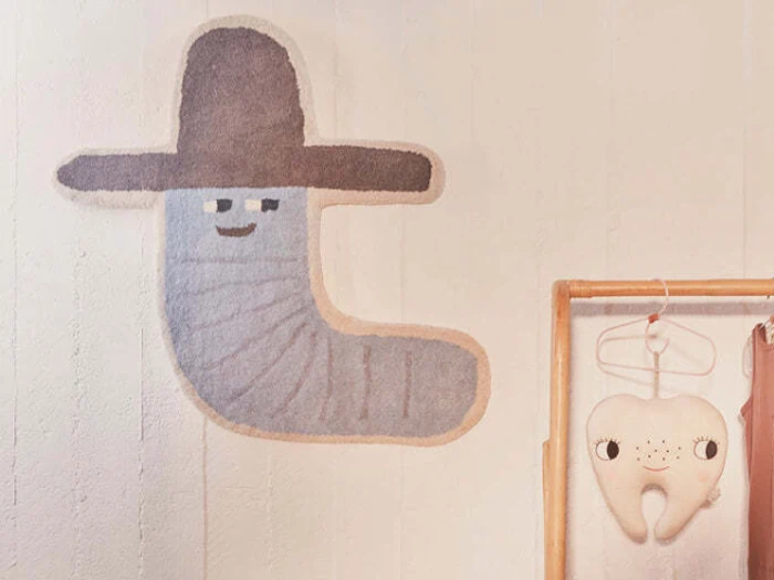 På en væg hænger en grå, ormelignende figur med ansigt og en brun hat. I nærheden hænger en plys-tand med ansigt på et træstativ.