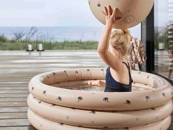 Et lille barn i en lille oppustelig pool holder en matchende oppustelig bold over hovedet på et trædæk nær vandet.