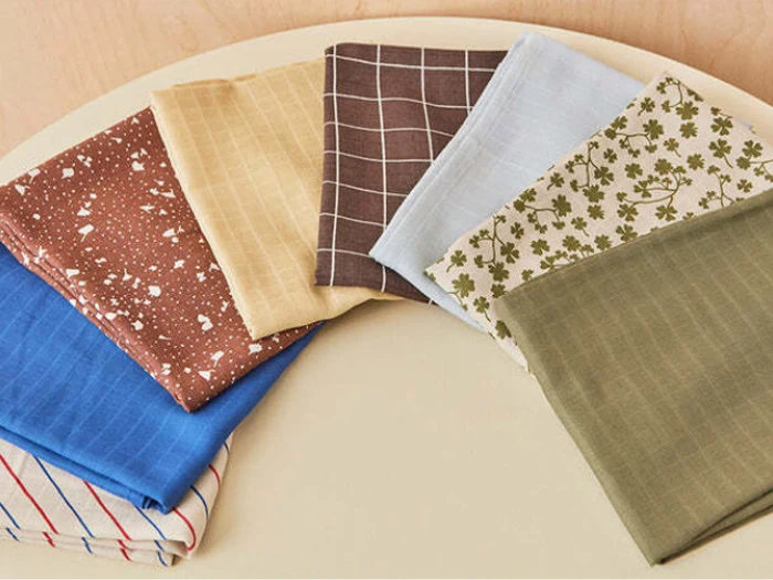 Seks stykker foldet stof med forskellige farver og mønstre, arrangeret i en halvcirkel på en lys overflade.