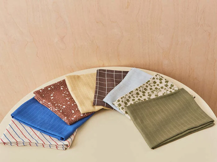 Syv foldede stofservietter i forskellige farver og mønstre er arrangeret i en halvcirkel på et lyst rundt bord mod en beige væg.