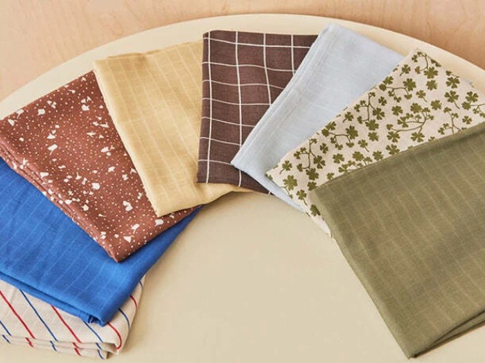 Seks foldede stoffirkanter i forskellige farver og mønstre, herunder striber, tern, blomster og plettede mønstre, arrangeret i en halvcirkel på en lys overflade.