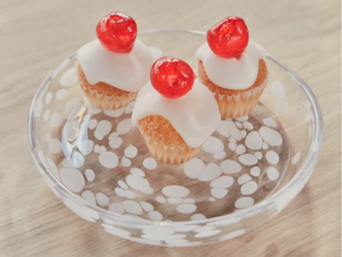 Tre små cupcakes med hvid glasur og et rødt kirsebær på toppen, arrangeret på en klar glastallerken med hvide polkaprikker, stillet på en lys træplade.