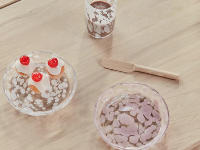 Et træbord med en klar glasplade med to små kager med kirsebær på toppen, en matchende glasskål, en træspatel og et glas chokolademælk.