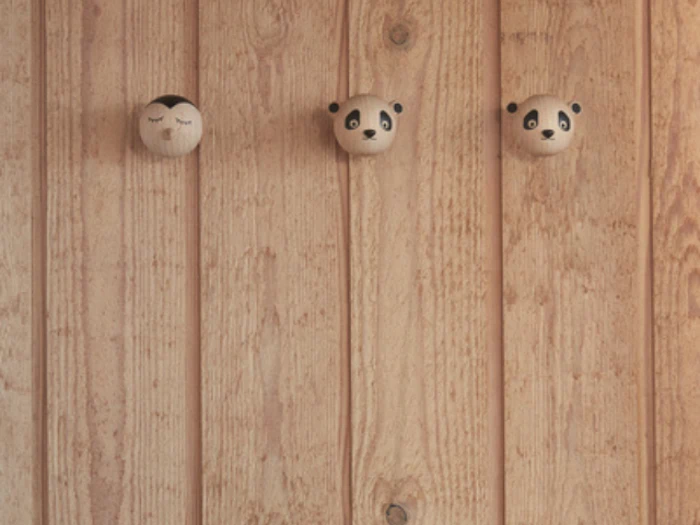 Tre vægkroge af træ på et træpanel; en har et ansigt med lukkede øjne, og to har pandabjørneansigter.