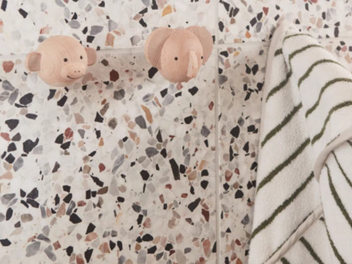 To dyreformede vægkroge af træ, den ene ligner en abe og den anden en elefant, er monteret på en plettet terrazzovæg og holder et stribet håndklæde.