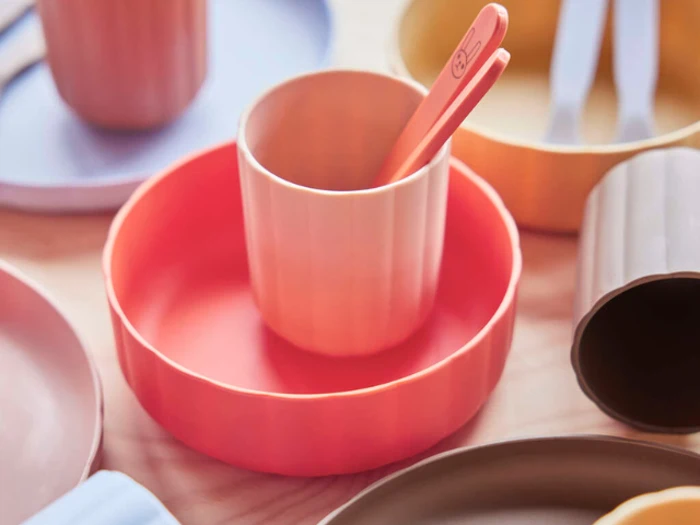 Farverige plastikkopper, tallerkener og skeer arrangeret på et bord, med en orange kop og matchende tallerken i midten af billedet.