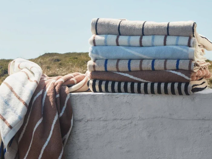 En stak foldede, stribede håndklæder i neutrale og blå toner ligger på en hvid væg udendørs med græsklædte bakker og blå himmel i baggrunden.