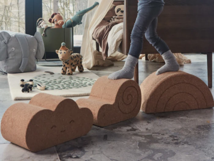 Et barn balancerer på legeklodser af kork formet som en sky, en spiral og en regnbue i et legerum med legetøj og et tæppe på gulvet.