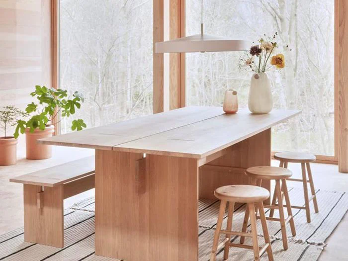 Et spisebord i lyst træ med taburetter og en bænk står ved store vinduer. Enkle vaser med blomster og potteplanter pynter i det minimalistiske, luftige rum.