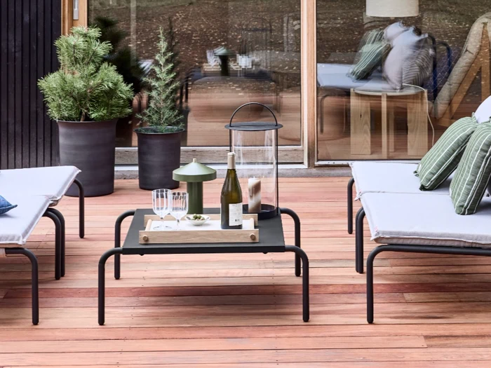 En udendørs terrasse med liggestole, et lavt bord med vin, glas, en lanterne og potteplanter; træterrasse og store glasdøre i baggrunden.