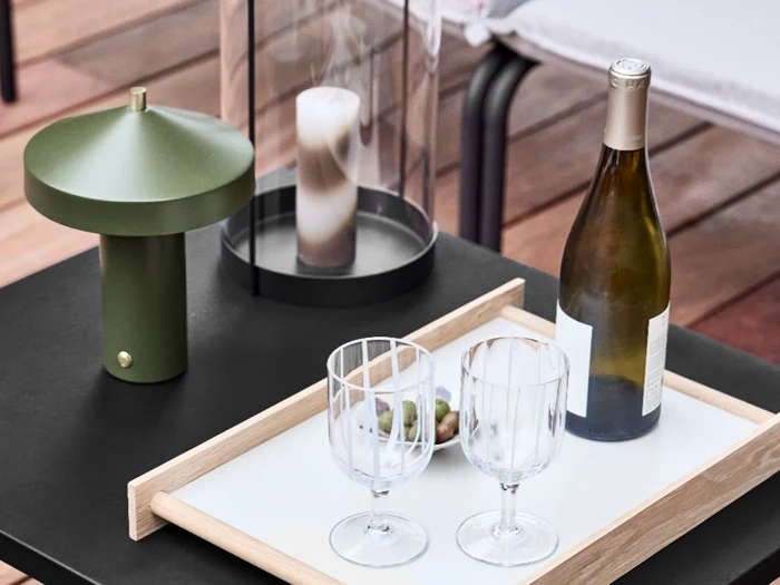 Et bord med en grøn lampe, et stearinlys i en glasholder, et bakke med en vinflaske, to tomme glas og en lille skål med oliven.