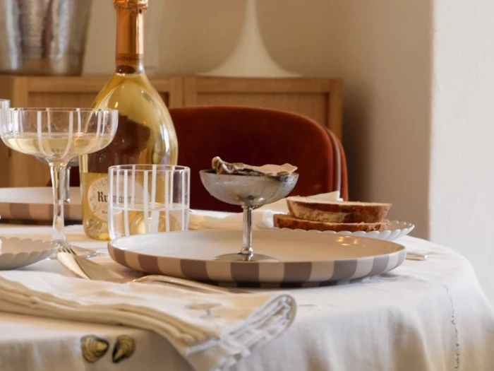 Et bord dækket med en stribet tallerken, et glas med østers, vinglas, en flaske mousserende vin og brødskiver på en hvid dug.