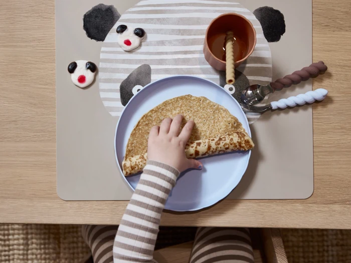 Et barn i stribet pyjamas rækker ud efter en sammenfoldet pandekage på en tallerken, mens der på bordet ligger en bordskåner med pandamotiv, en kop, bestik og kager formet som pandahoveder.