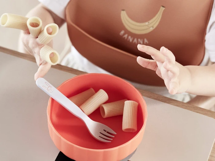 Et barn, der har en hagesmæk med bananmotiv på, har rigatoni-pasta på fingrene og rækker ud mod en skål med mere pasta og en gaffel.