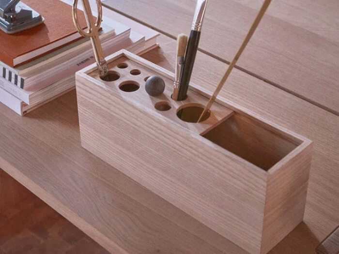 På et træbord ved siden af stablede notesbøger står en skrivebordsarrangør i træ med sakse, pensler og blyanter.