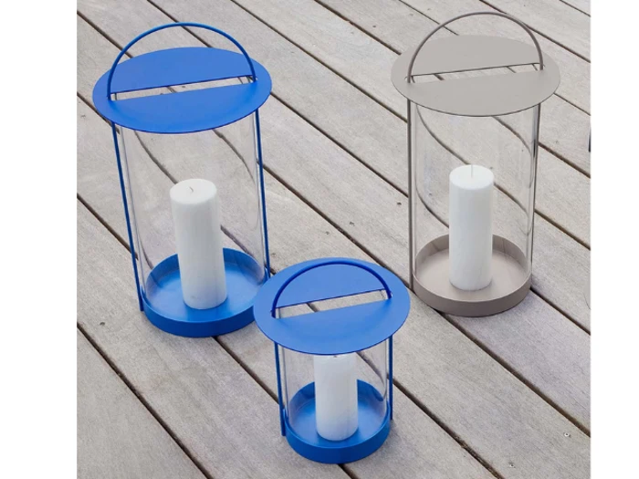 Tre cylindriske lanterner med håndtag, to blå og en grå, hver med et hvidt bloklys, placeret på et lyst trædæk.