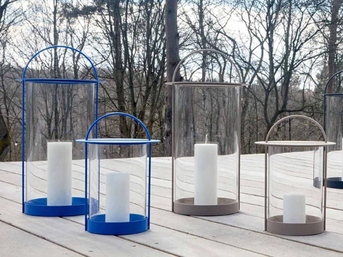 Fire glaslanterner med hvide bloklys indeni er placeret på et trædæk med bladløse træer i baggrunden.