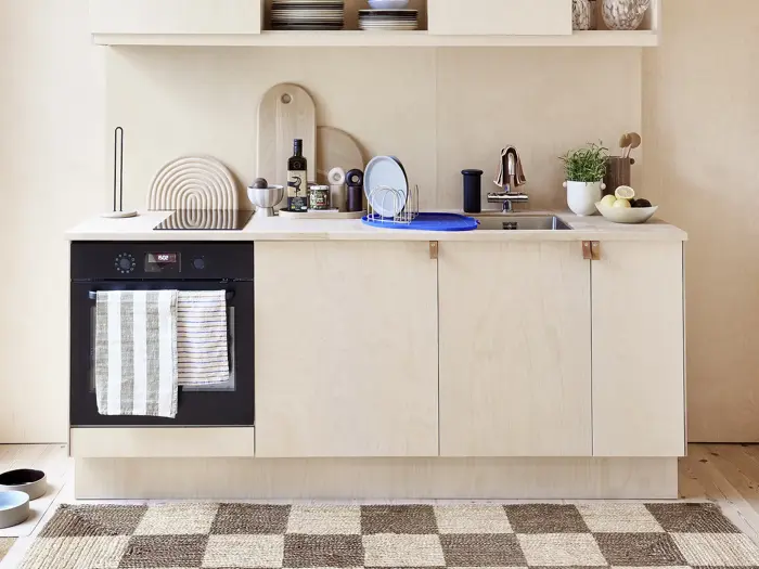 Et moderne køkken i lyst træ med indbygget ovn, stribet håndklæde, vask, skærebrætter, redskaber, planter og pænt arrangeret service på køkkenbordet og hylderne ovenover.