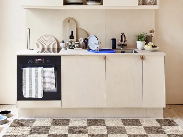 Minimalistisk køkken i lyst træ med ovn, stribede håndklæder, forskellige redskaber, skærebrætter, en potteplante og tallerkener på køkkenbordet og et ternet tæppe på gulvet.