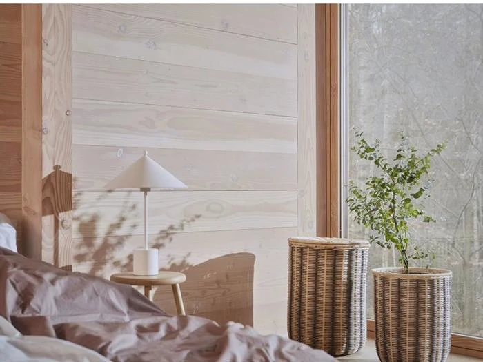 Et hyggeligt værelse med lyse trævægge, et natbord med en hvid lampe, to flettede kurve, en plante og et stort vindue med træer udenfor.