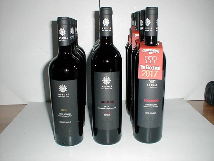 Ni flasker Assuli-rødvin er anbragt i rækker på en hvid flade; en flaske har en rød etiket med teksten Tre Bicchieri 2017.