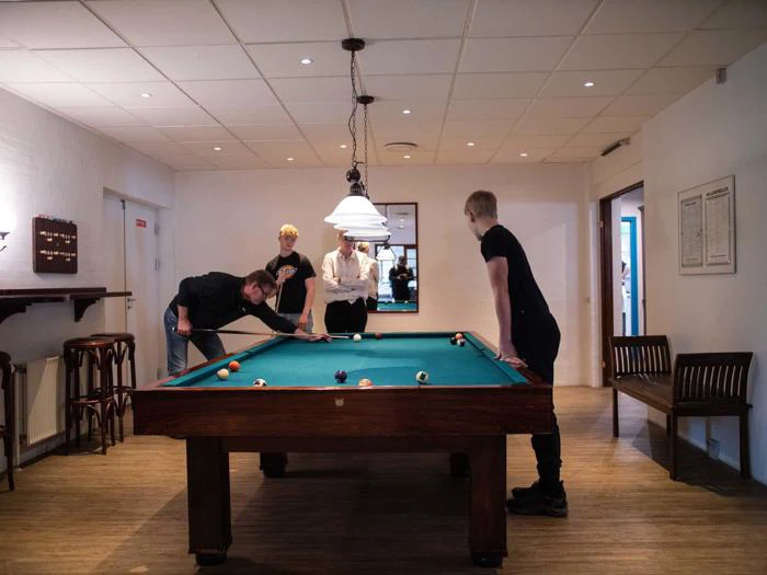 Fire unge mænd spiller pool i et veloplyst rum med trægulv; en tager et slag, mens de andre ser til rundt om bordet.