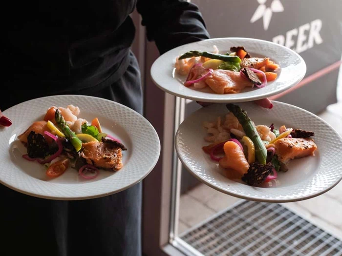 En person holder tre hvide tallerkener, hver med en gourmetret med laks, asparges, cherrytomater og syltede løg.