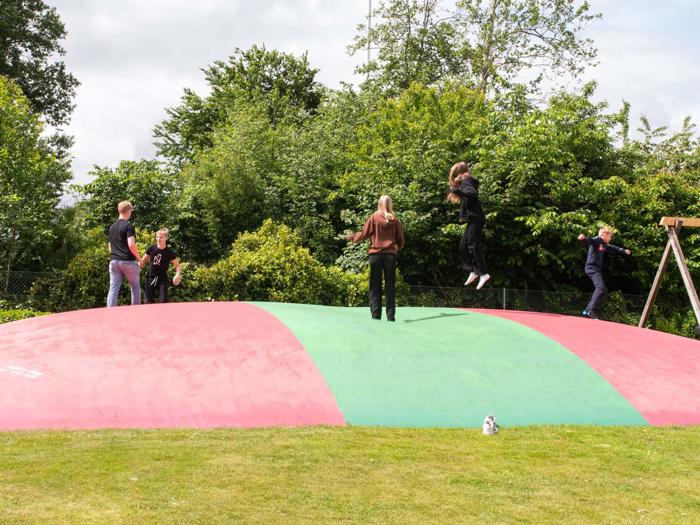 Fem personer leger og hopper på en stor, rød og grøn oppustelig legepude udendørs, omgivet af træer og græs.