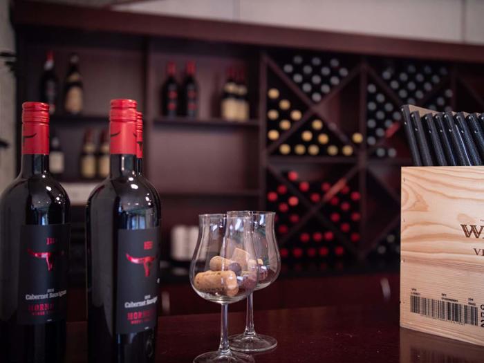 To flasker rødvin, to glas med propper og en trææske med kuglepenne på en disk med hylder med vinflasker i baggrunden.