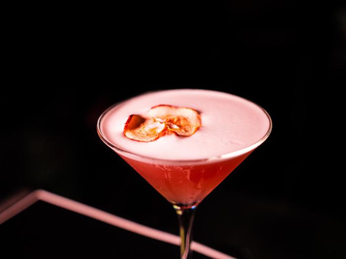 En lyserød cocktail i et martini-glas med en skummende top, pyntet med to tynde æbleskiver, på en mørk baggrund.